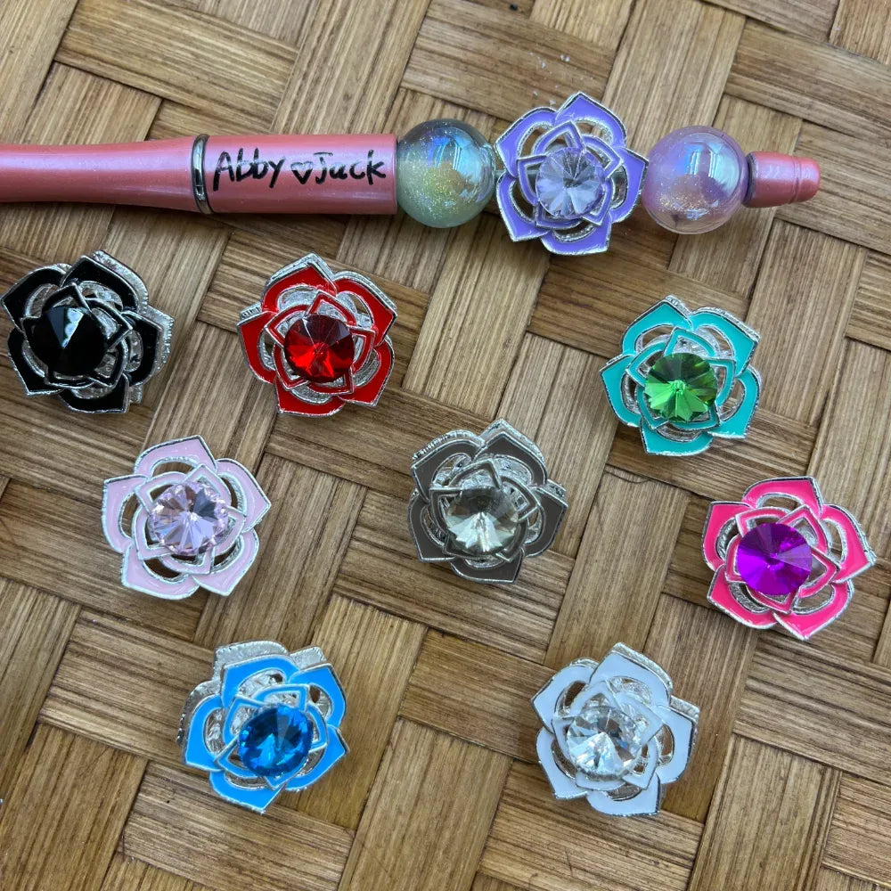 Assorted rose flower enamel metal beads with rhinestone crystal centers in multiple colors including black, red, pink, gray, teal, hot pink, blue, white, and purple, displayed on a wicker surface alongside a beadable pen.