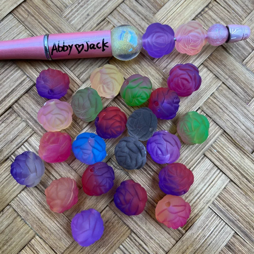 Multicolor frosted rose flower shaped acrylic beads in purple, pink, red, green, blue and ombre color combinations displayed on woven surface next to a beadable pen labeled Abby and Jack.