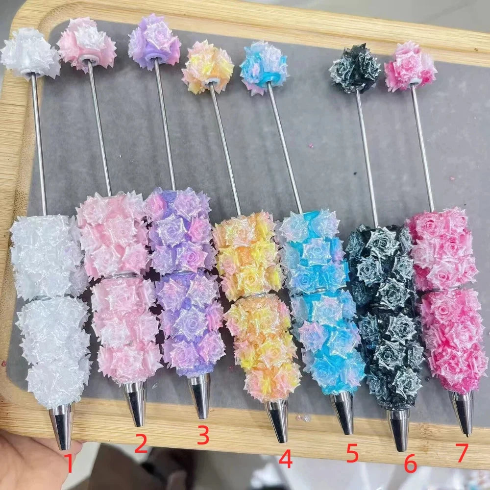 Seven glitter rose flower clay beadable pens in assorted colors including white, pink, lavender, yellow, blue, black, and hot pink displayed on a wooden board at DIY Mini Planet.