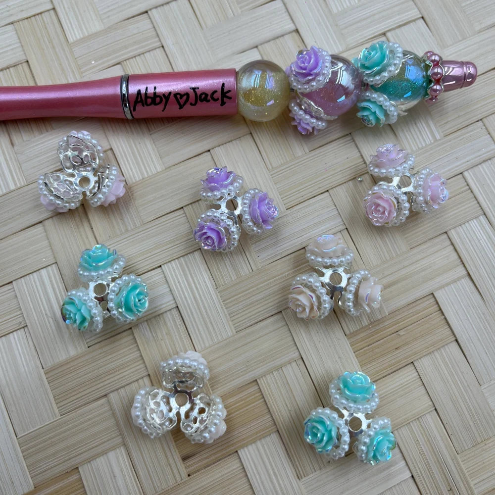 Decorative rose flower bead caps with pearl trim in mint green, purple, and pink pastel colors displayed on woven bamboo background next to a pink beadable pen labeled Abby Jack.