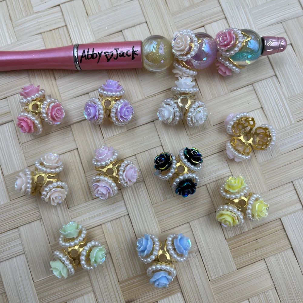 Assorted color resin rose flower bead caps with pearl rim and gold base, displayed on woven bamboo surface alongside a decorated beadable pen, available in pink, purple, white, black, yellow, green, and blue.