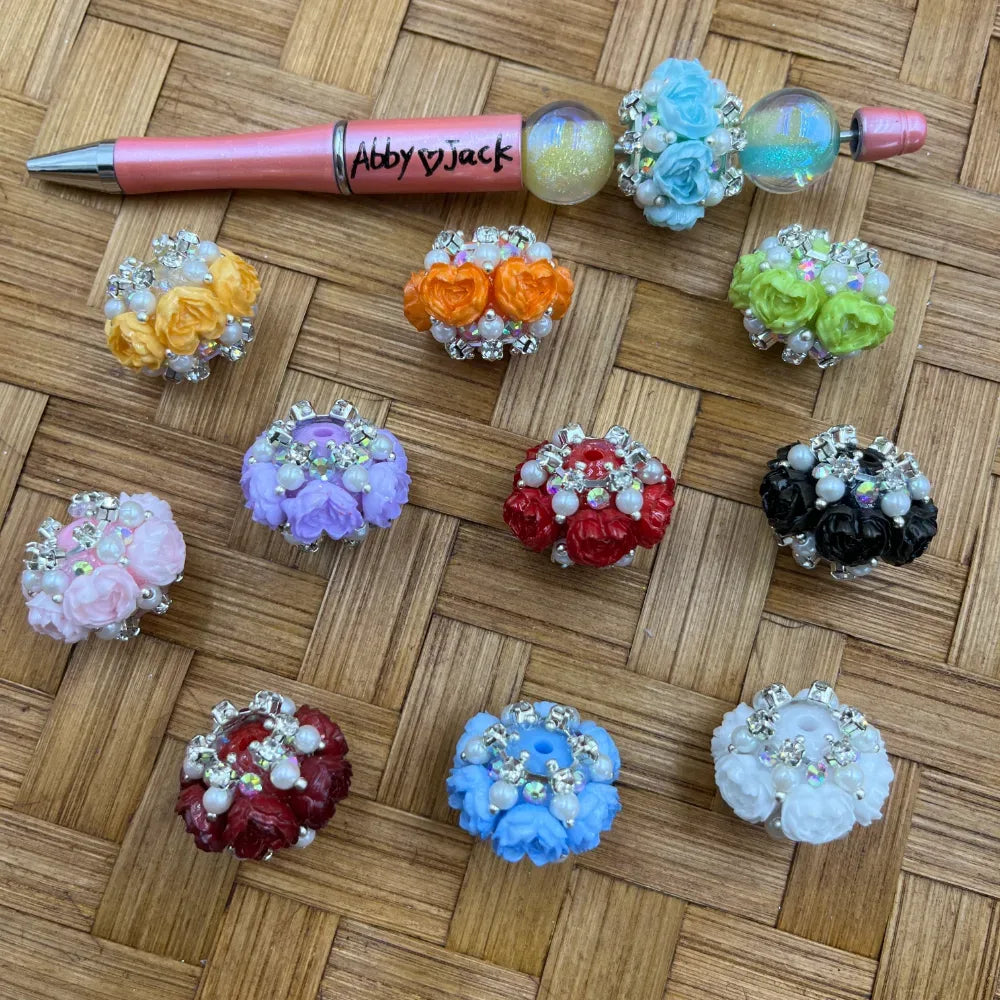 Assorted rose flower fancy beads with rhinestones and pearl accents in 10 colors including pink, red, orange, yellow, purple, green, blue, black, white and dark red, displayed on wicker surface next to a decorated beadable pen.