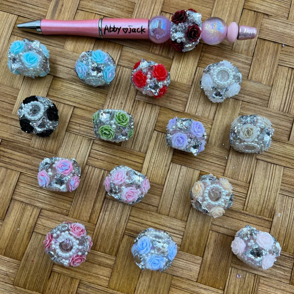 Assorted fancy beads decorated with colorful miniature roses, rhinestones, pearl accents, and silver foil in multiple colors including pink, red, blue, purple, green, black, and white, displayed on a woven surface alongside a decorated beadable pen.