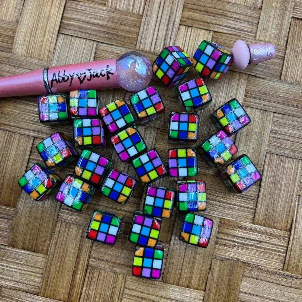 Colorful Rubik's Cube patterned acrylic cube beads scattered on a woven surface next to a pink beadable pen, showing mixed multicolor grid designs.