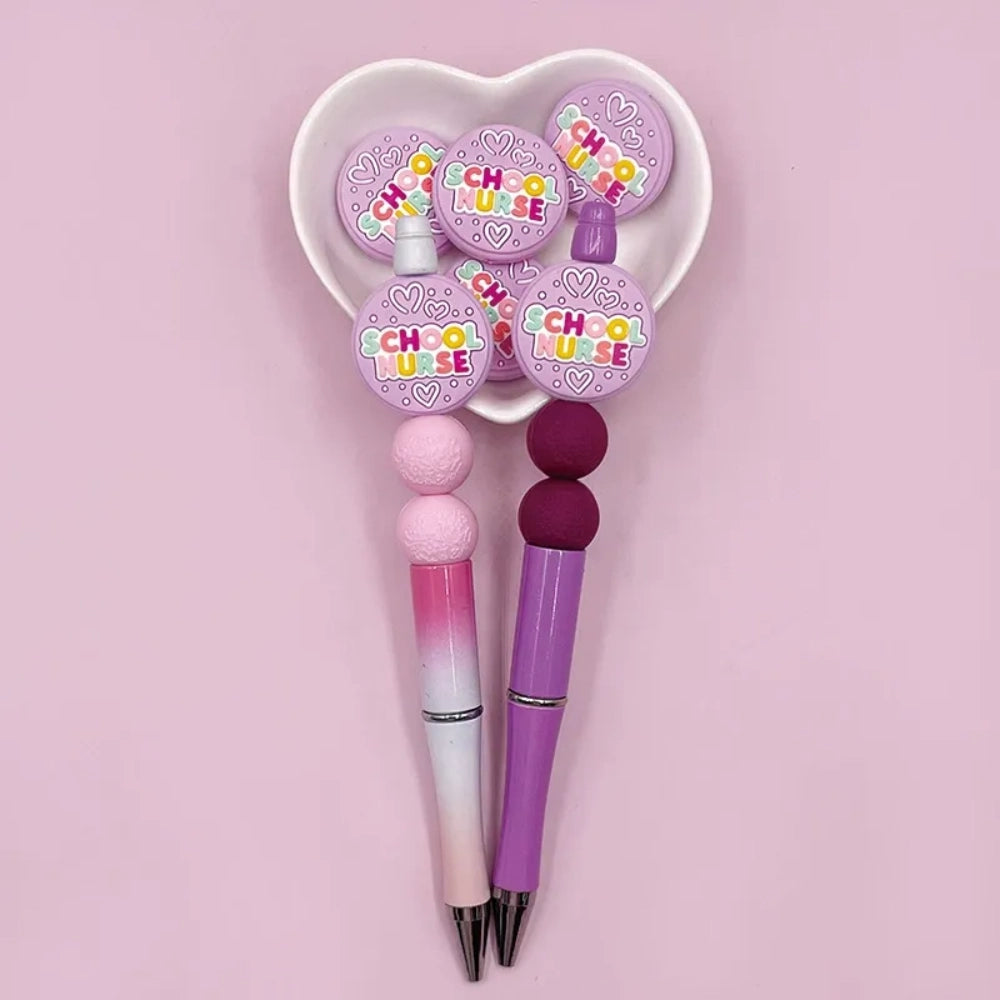 Colorful School Nurse silicone focal beads in purple with multicolor lettering and heart details, shown on beaded pens and in a heart-shaped dish on a pink background.