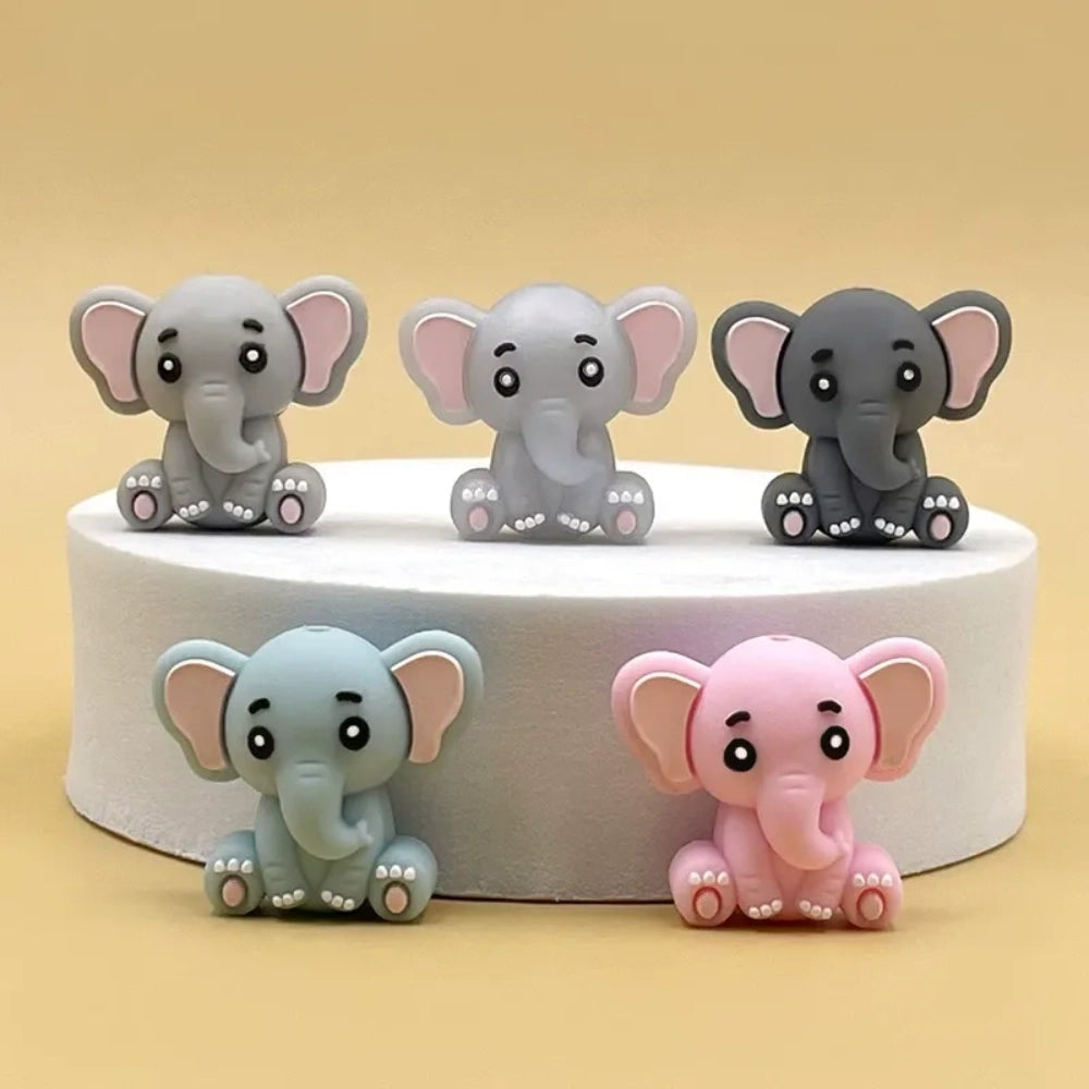 Five cute 3D baby elephant silicone focal beads in light gray, medium gray, dark gray, blue, and pink colors, sitting posed with pink inner ears and toenails, displayed on a white platform against a warm beige background.