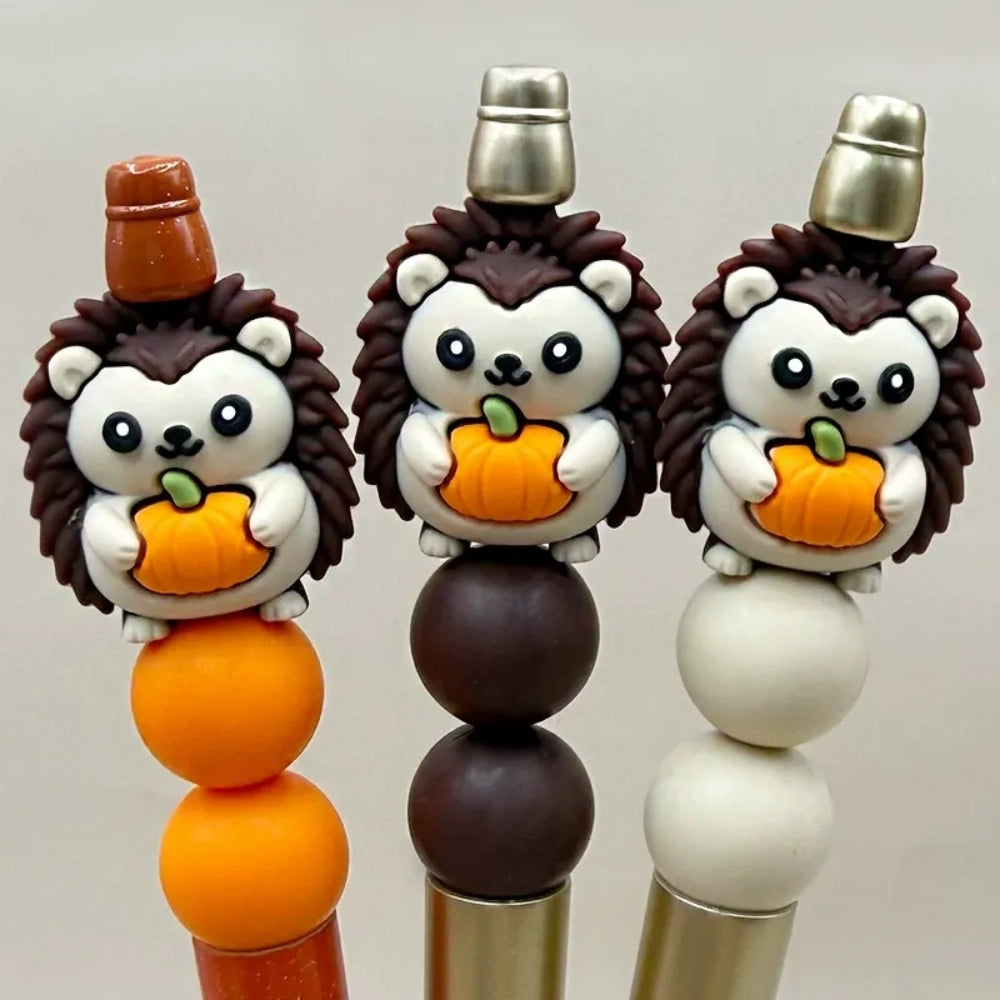 Three 3D silicone hedgehog focal beads holding orange pumpkins, shown on beaded pens in orange, brown, and cream colorways, perfect for fall beadable pen and keychain crafts.