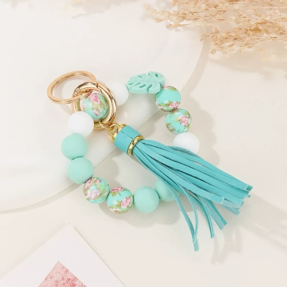 Two handmade silicone bead keychains with leather tassels — one in teal with floral print beads and one in sage green with leopard print and monstera leaf silicone beads, displayed on a white marble surface. - Blue