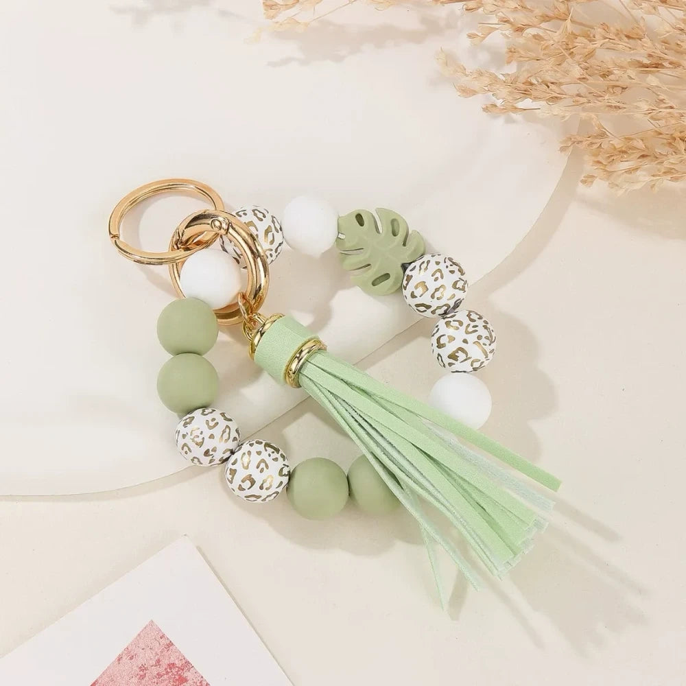 Two handmade silicone bead keychains with leather tassels — one in teal with floral print beads and one in sage green with leopard print and monstera leaf silicone beads, displayed on a white marble surface. - Green