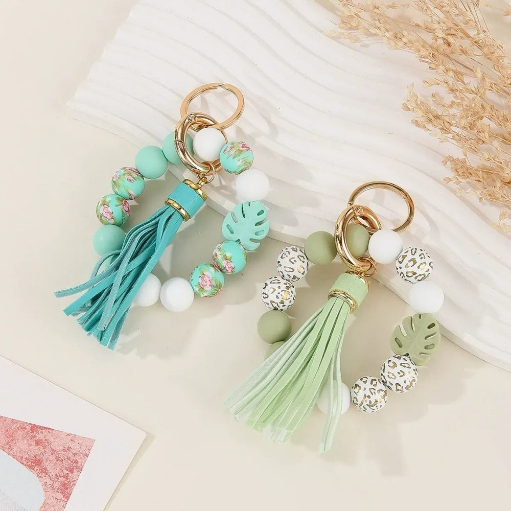 Two handmade silicone bead keychains with leather tassels — one in teal with floral print beads and one in sage green with leopard print and monstera leaf silicone beads, displayed on a white marble surface. - Random Mixed