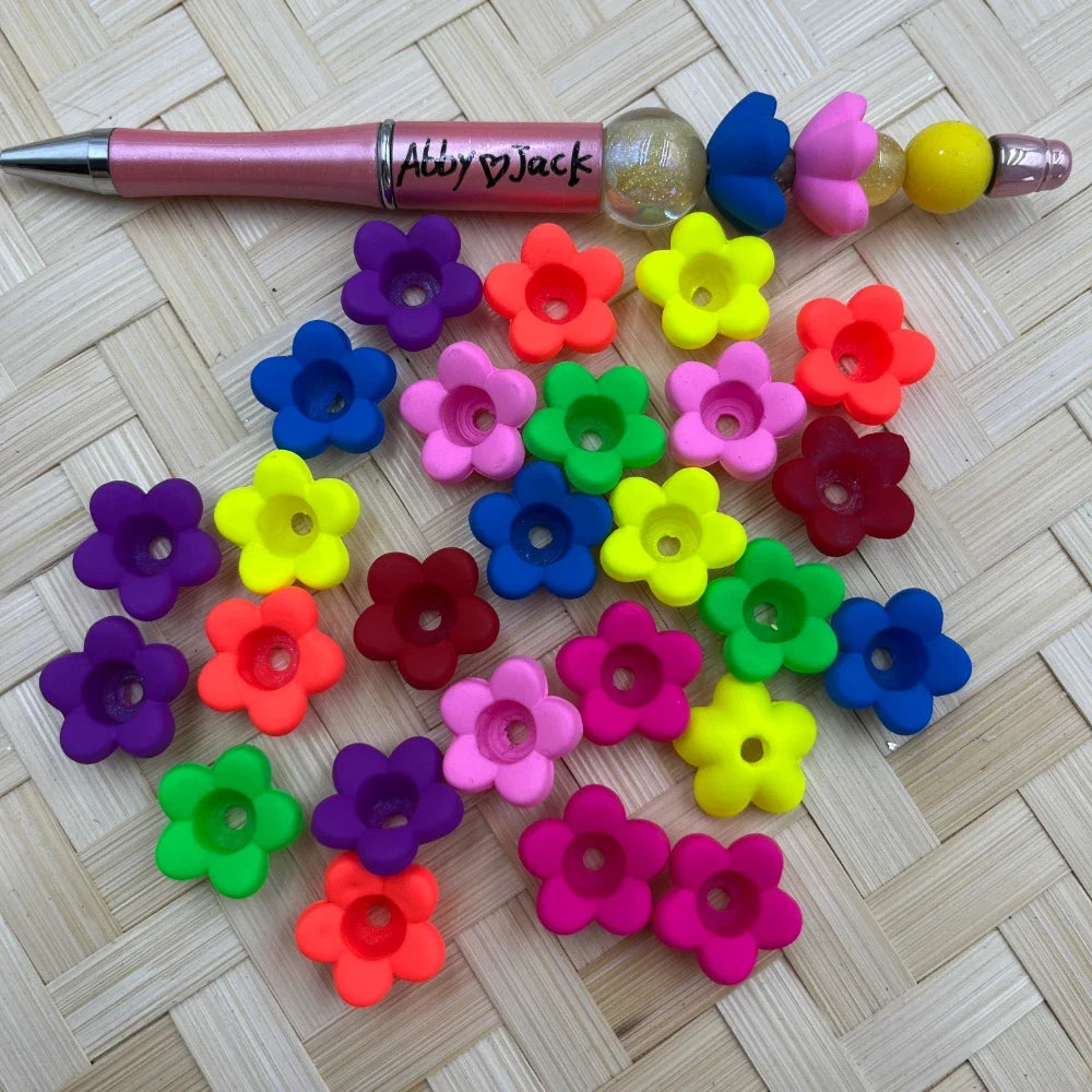 Assorted colorful silicone flower-shaped bead caps in neon and bright mixed colors displayed on a woven bamboo surface next to a decorated beadable pen.