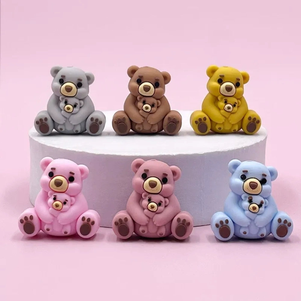 Six colorful 3D silicone focal beads shaped like a mama bear hugging a baby bear, available in gray, brown, yellow, pink, dusty pink, and blue, displayed on a white pedestal with a pink background. - Random Mixed