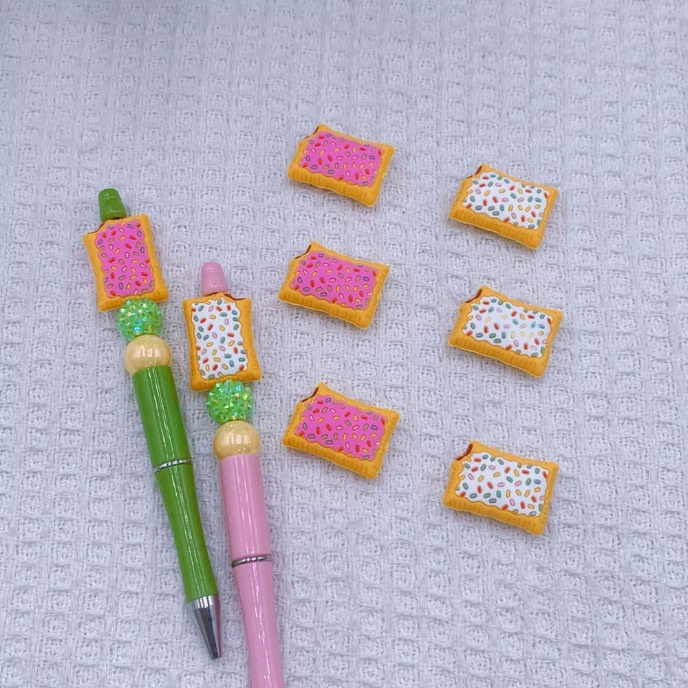 Miniature 3D Pop Tart silicone focal beads with pink and white frosting and colorful sprinkles, shown on green and pink beadable pens alongside loose focal beads on a white waffle texture background. - Random Mixed