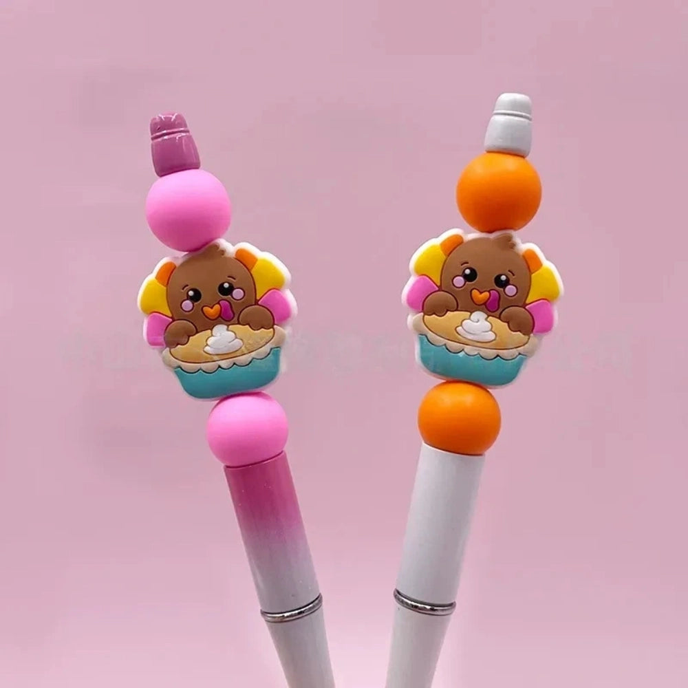 Two beadable pens decorated with cute 3D turkey cupcake silicone focal beads in pink and orange colorways on a pink background, perfect for DIY crafting. - Random Mixed