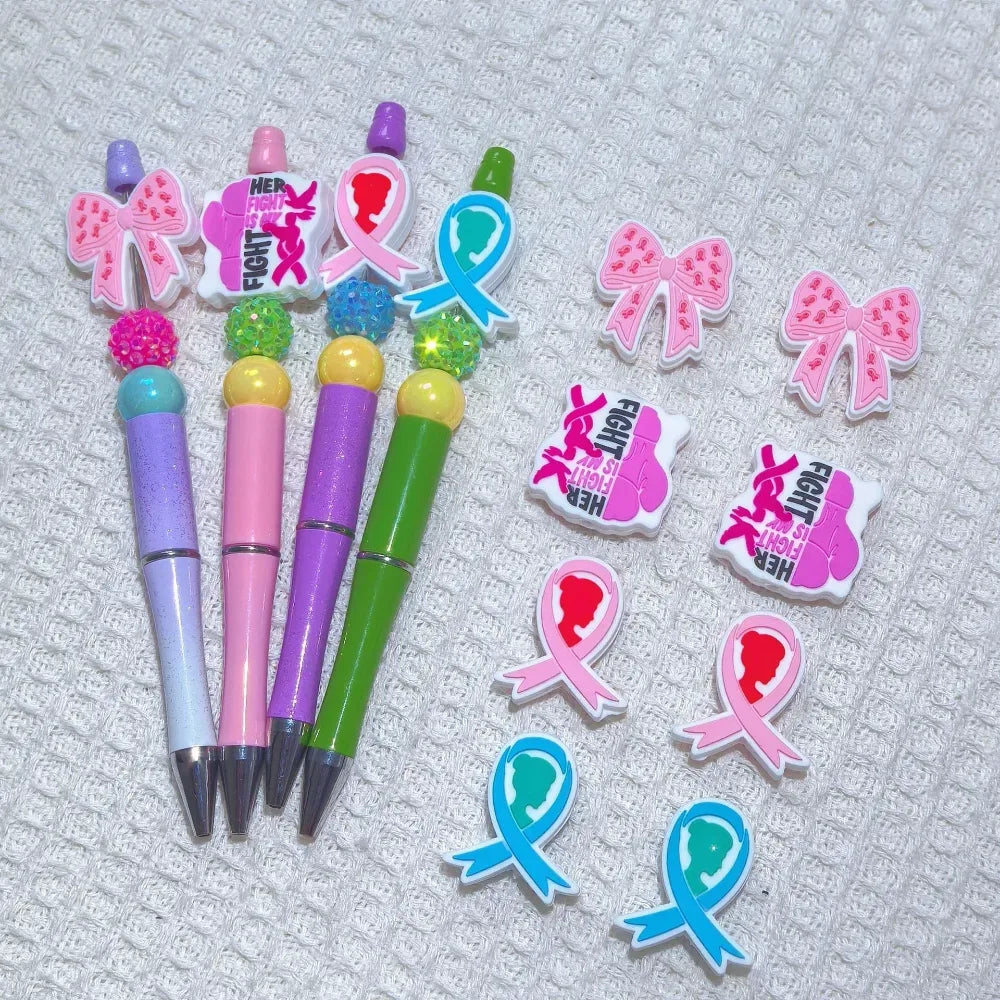 Silicone 3D focal beads in cancer awareness designs including pink and blue awareness ribbons, decorative bows, and 'Her Fight Is My Fight' charms displayed alongside completed beadable pens on a white textured background.