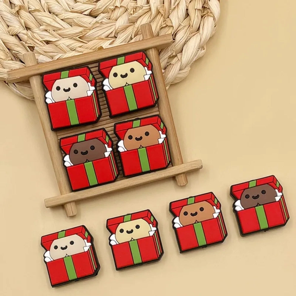 Cute silicone focal beads shaped like red and green Christmas gift boxes with smiling character faces peeking out, shown in multiple skin tone variations on a wooden display tray. - Random Mixed