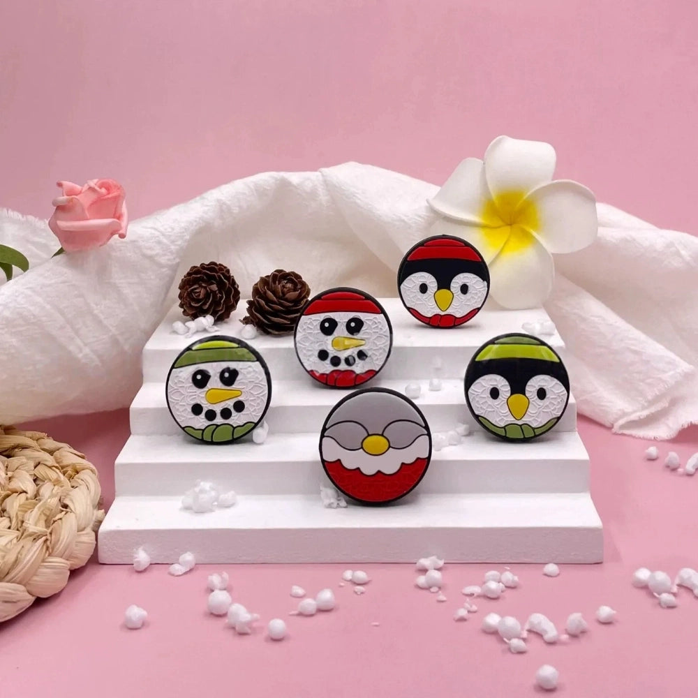 Six round Christmas silicone focal beads featuring snowman and penguin designs with red and green hats displayed on a white tiered stand with holiday decorations on a pink background.