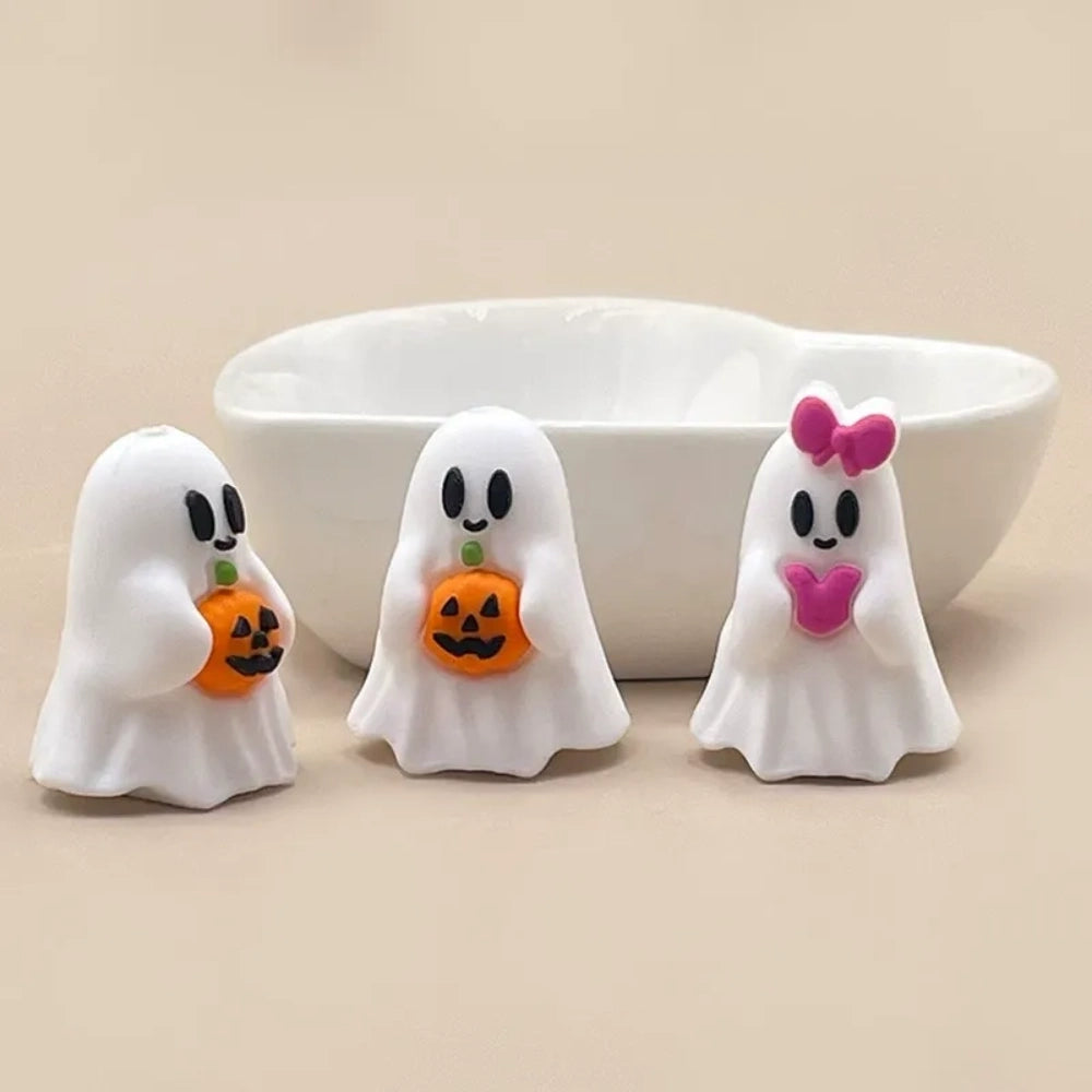 Three white Halloween ghost silicone focal beads – two holding jack-o-lantern pumpkins and one with a pink bow and heart – displayed in front of a white ceramic bowl on a beige surface.
