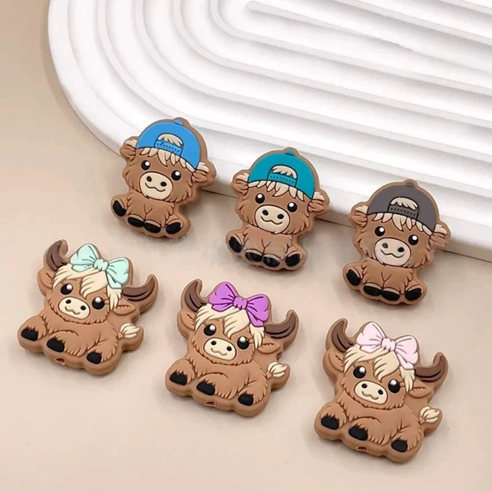 Six silicone Highland cow focal beads in brown – three wearing colorful baseball caps (blue, teal, gray) and three with bow accessories (mint, purple, pink) – crafting supplies for beadable pens, keychains, and badge reels. - Random Mixed