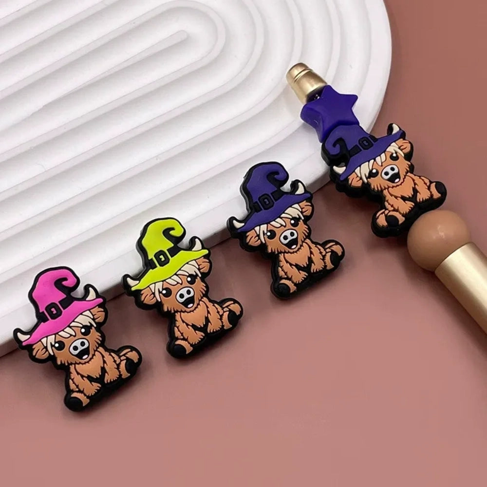 Four 3D silicone focal beads shaped like cartoon highland cows wearing witch hats in pink, yellow-green, purple, and dark purple, displayed on a white tray with a beadable pen. - Random Mixed