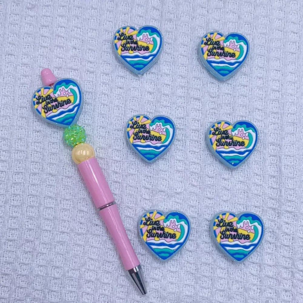 Silicone focal beads featuring Jesus Loves You round, Best Mom Ever star, and Live in the Sunshine heart designs displayed on beadable pens and scattered on a white plate, colorful DIY craft supplies from DIY Mini Planet. - 1