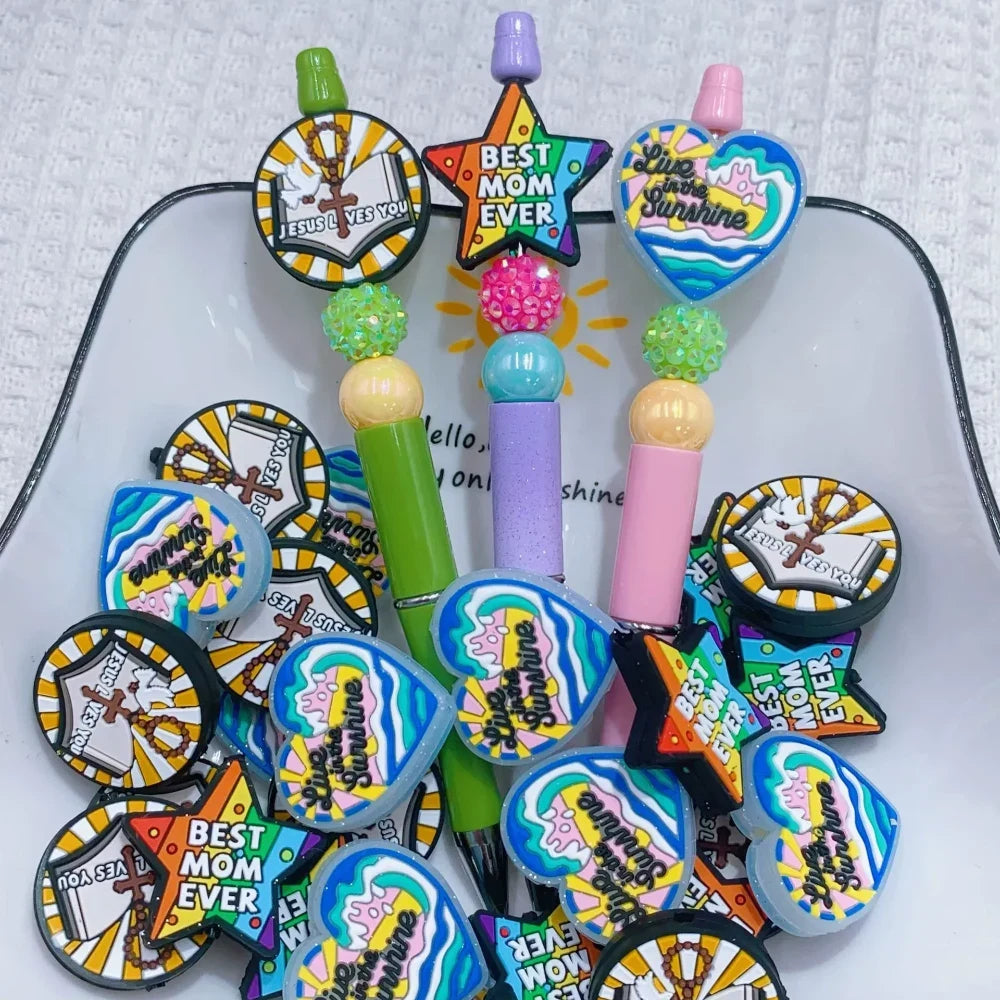 Silicone focal beads featuring Jesus Loves You round, Best Mom Ever star, and Live in the Sunshine heart designs displayed on beadable pens and scattered on a white plate, colorful DIY craft supplies from DIY Mini Planet. - Random Mixed