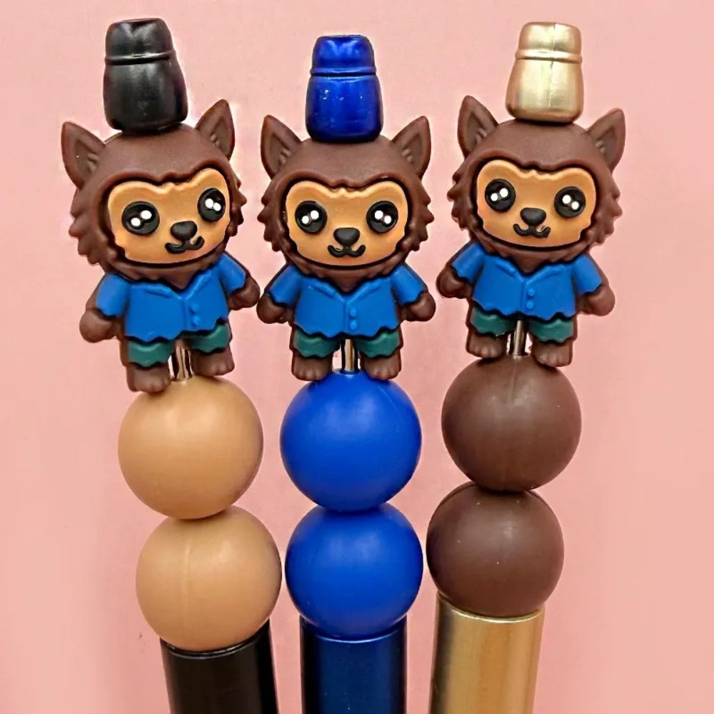 Three kawaii werewolf silicone focal beads wearing blue shirts and teal pants, shown on beadable pens with tan, blue, and brown silicone beads against a pink background.