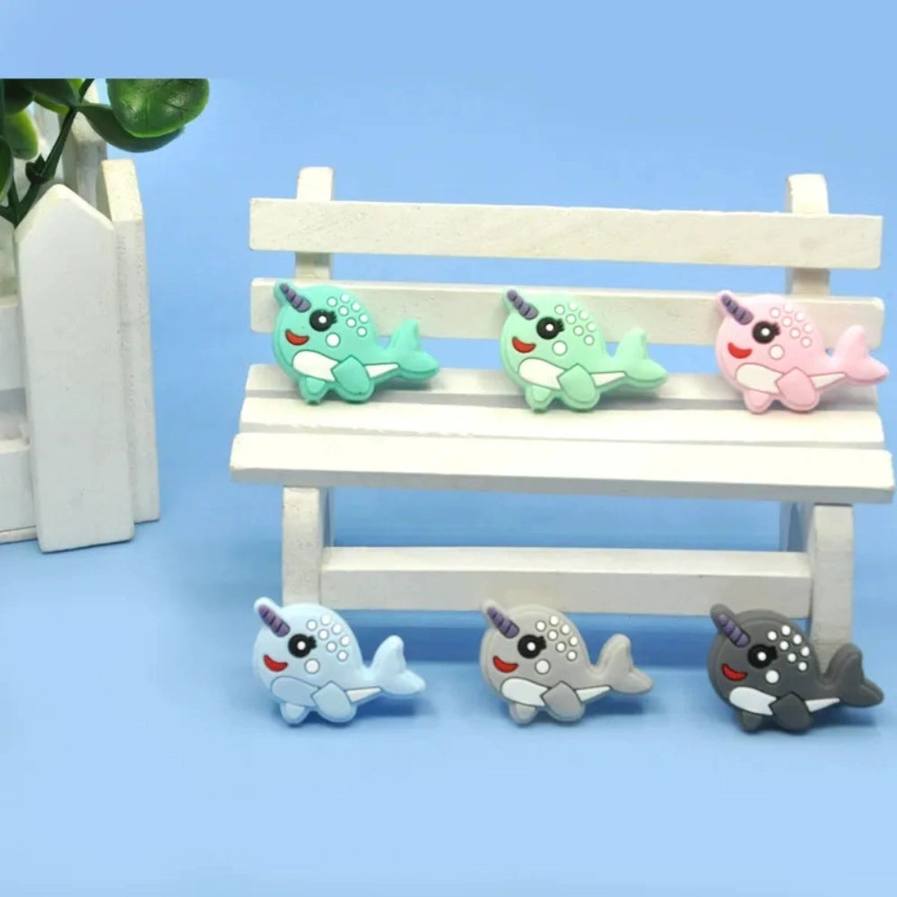 Set of 5 kawaii narwhal silicone focal beads in teal, mint, pink, light blue, gray, and dark gray, displayed on a miniature white bench with blue background.
