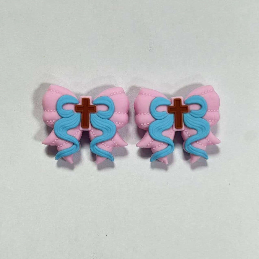 Two pink bow silicone focal beads with blue ribbon swirls and brown cross detail, shown side by side on white background, for beadable pens and DIY crafts.