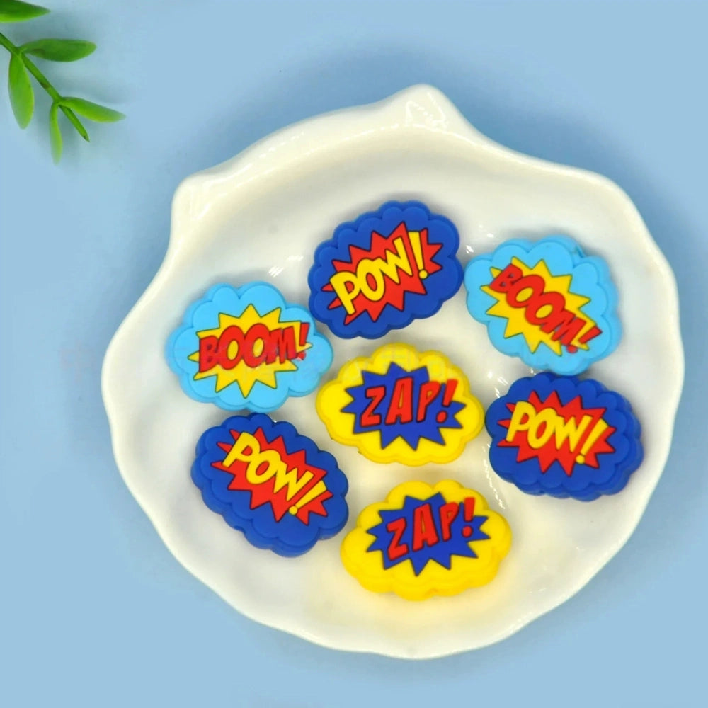 Colorful POW, BOOM, and ZAP superhero comic book silicone focal beads in blue, yellow, and light blue displayed on a white shell dish, perfect for beadable pens, keychains, and badge reels. - Random Mixed