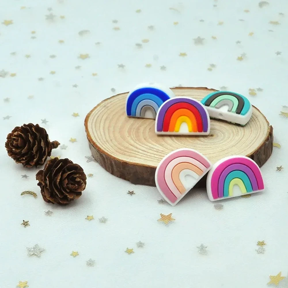 Five colorful silicone rainbow arch focal beads in various color combinations including blue-gray, multicolor, dark-mint, boho pink, and pink-teal, displayed on a wooden slice with pine cones on a star-patterned surface.