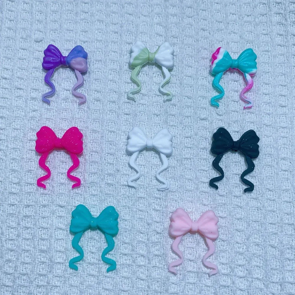 8 assorted ribbon bow silicone focal beads in purple, mint, teal swirl, hot pink, white, black glitter, turquoise, and light pink, displayed on white waffle fabric background.
