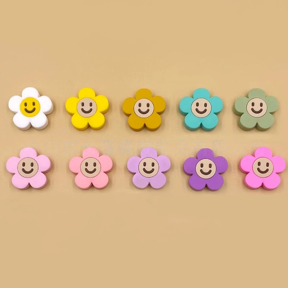 10 colorful smiley face daisy flower silicone focal beads in white, yellow, mustard, teal, sage, pink, light pink, lavender, purple, and hot pink on a tan background.