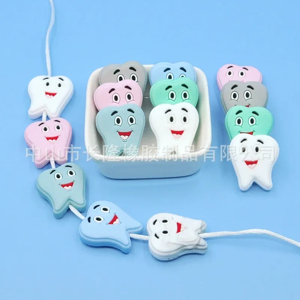 Assorted pastel smiling tooth shaped silicone focal beads in white, pink, blue, green, and gray with cartoon faces, strung on cord and displayed in a white tray on a blue background.
