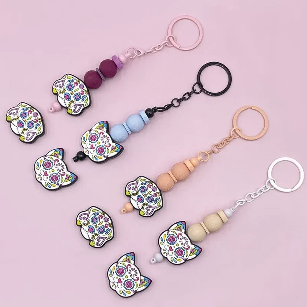 Sugar skull cat silicone focal beads in colorful Day of the Dead floral design, shown assembled on beaded keychains with pink, blue, tan, and natural wood beads on a pink background.