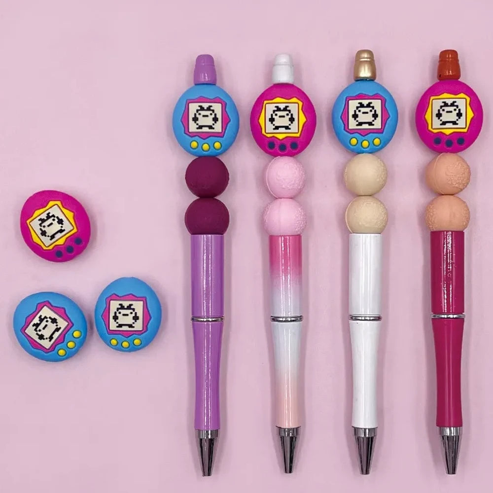 Assorted Tamagotchi silicone focal beads in pink and blue colorways displayed alongside four completed beadable pens on a pink background, showing pixel pet virtual pet screen design detail.