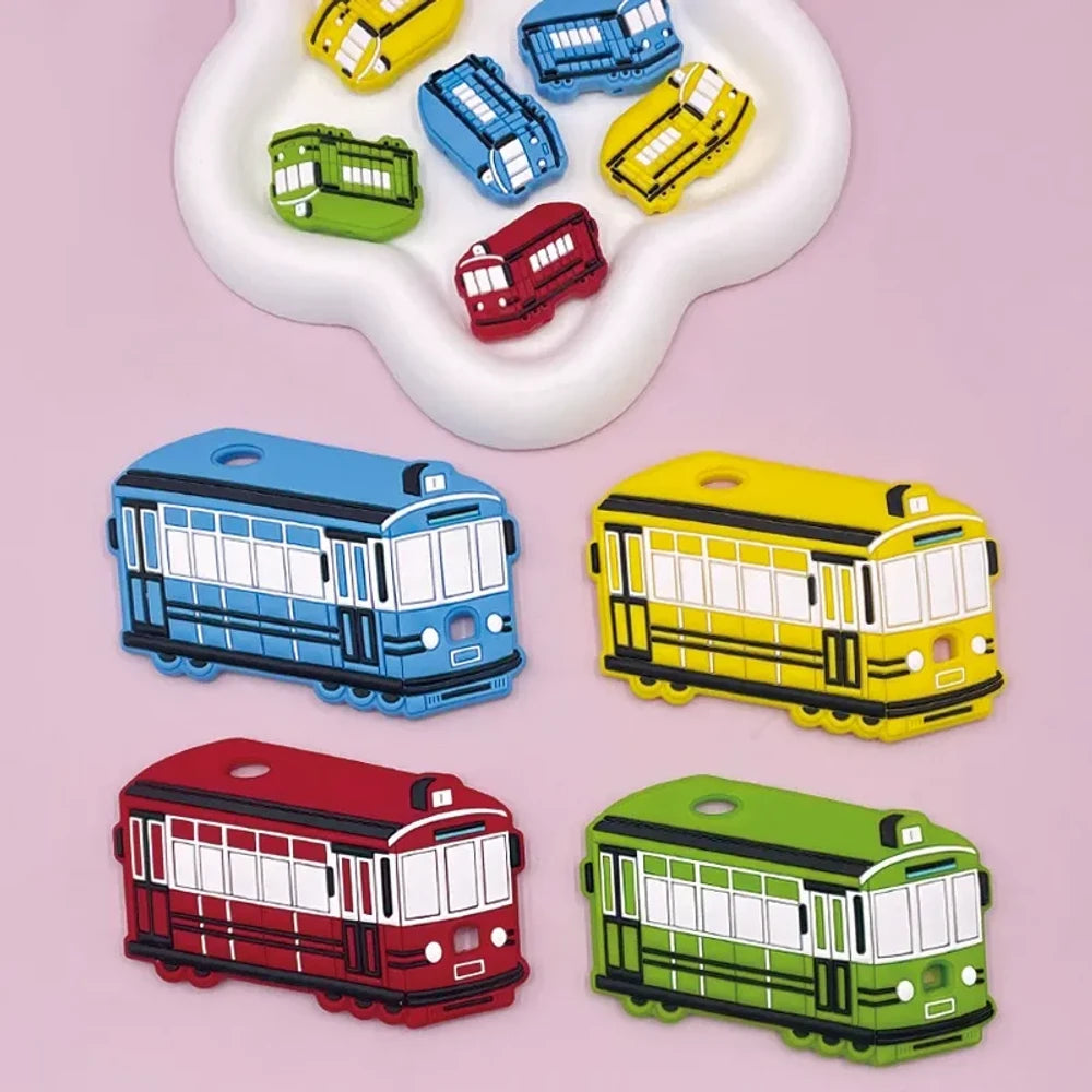 Four colorful silicone tram streetcar focal beads in blue, yellow, red, and green displayed on a pink background with miniature versions shown in a white dish above.