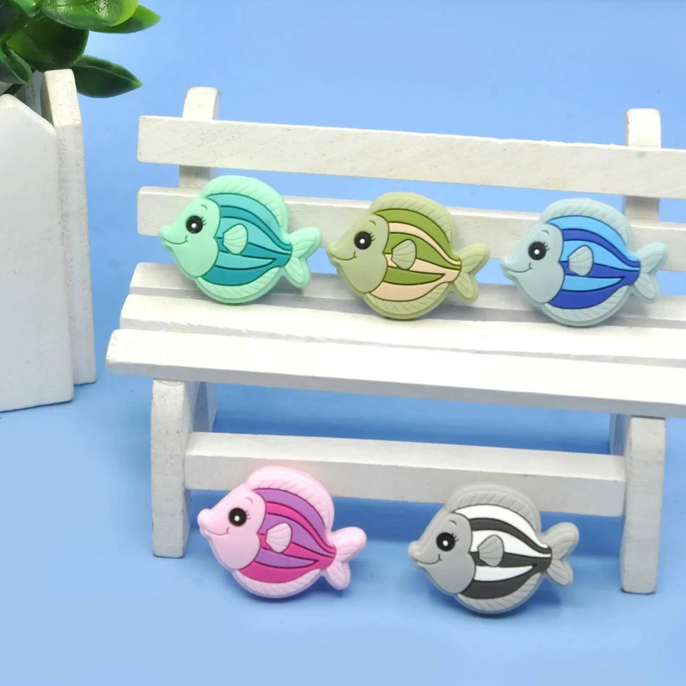Five colorful cartoon tropical fish silicone focal beads in teal, olive green, blue, pink, and gray displayed on a miniature white wooden bench against a blue background.
