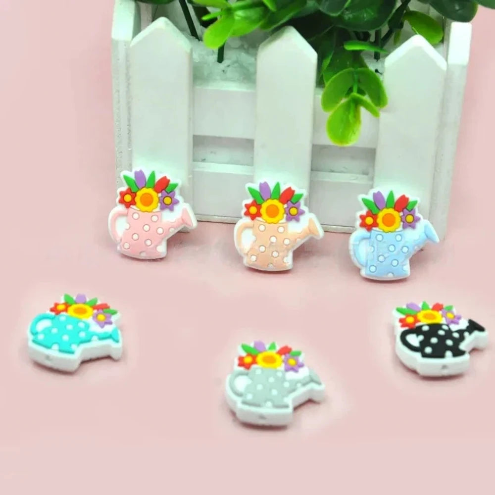 Five colorful silicone focal beads shaped like polka dot watering cans with 3D flower bouquets on top, displayed in pink, peach, blue, teal, white, and black colorways on a pink surface with a white picket fence backdrop.