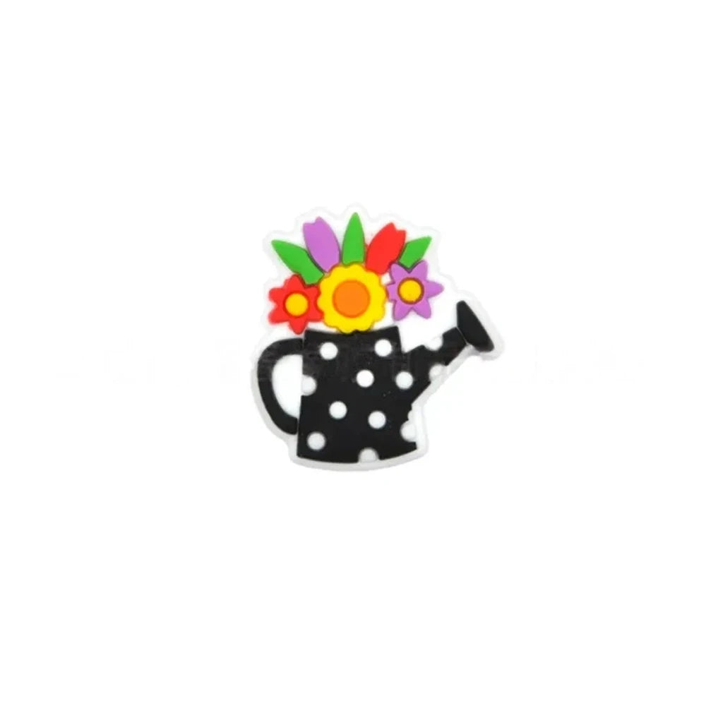 Five colorful silicone focal beads shaped like polka dot watering cans with 3D flower bouquets on top, displayed in pink, peach, blue, teal, white, and black colorways on a pink surface with a white picket fence backdrop. - Black