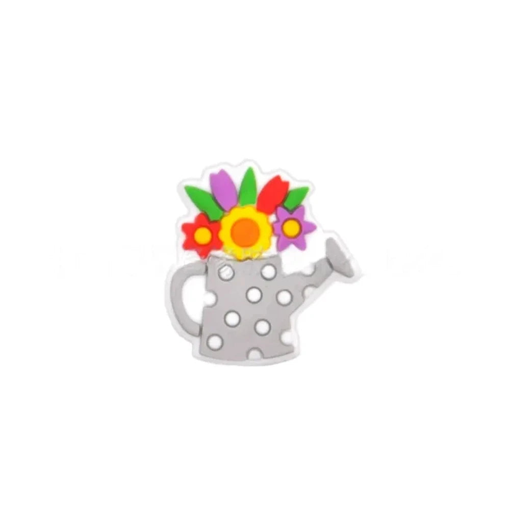 Five colorful silicone focal beads shaped like polka dot watering cans with 3D flower bouquets on top, displayed in pink, peach, blue, teal, white, and black colorways on a pink surface with a white picket fence backdrop. - Light Gray