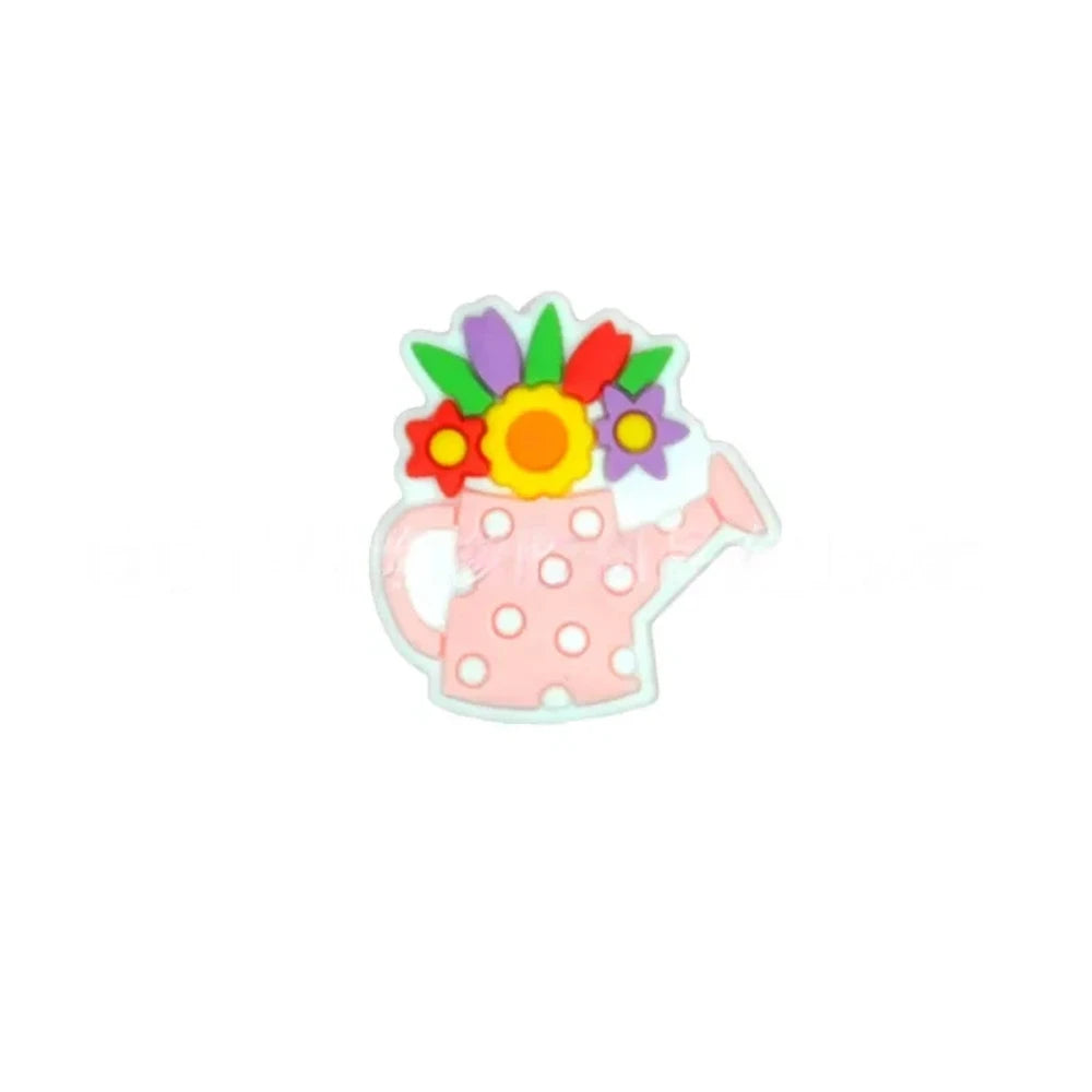 Five colorful silicone focal beads shaped like polka dot watering cans with 3D flower bouquets on top, displayed in pink, peach, blue, teal, white, and black colorways on a pink surface with a white picket fence backdrop. - Pink
