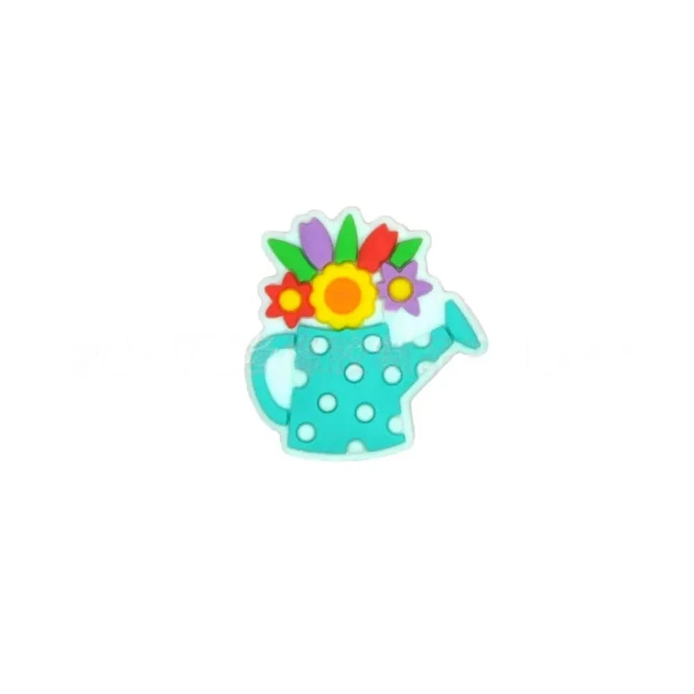 Five colorful silicone focal beads shaped like polka dot watering cans with 3D flower bouquets on top, displayed in pink, peach, blue, teal, white, and black colorways on a pink surface with a white picket fence backdrop. - Turquoise