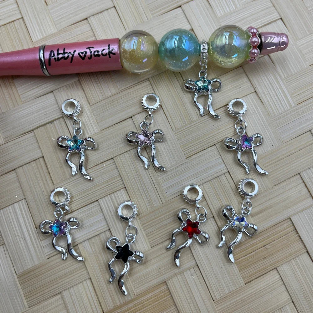 Silver metal bow ribbon charm bail beads with assorted colored star crystal centers in teal, pink, purple, black, red, and iridescent, displayed next to a beaded beadable pen on a wicker surface.