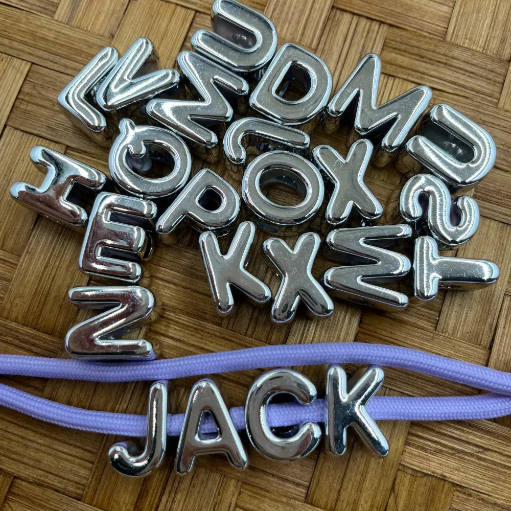 Silver chrome metallic alphabet letter acrylic beads A-Z scattered on bamboo surface with purple cord spelling JACK, showing individual letter beads for beadable pens and keychains.