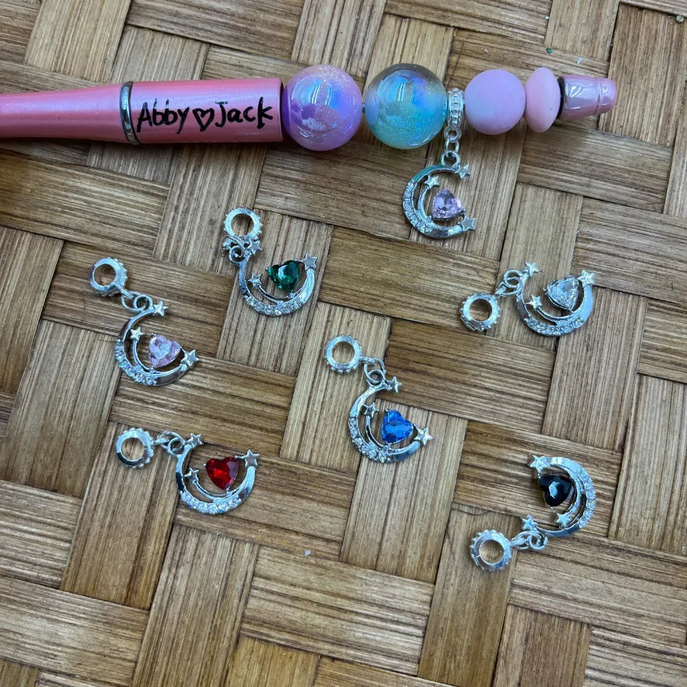 Assorted silver crescent moon charm bail beads with heart-shaped crystals in pink, green, red, blue, black, and clear gemstones displayed on a wicker surface next to a decorated beadable pen.