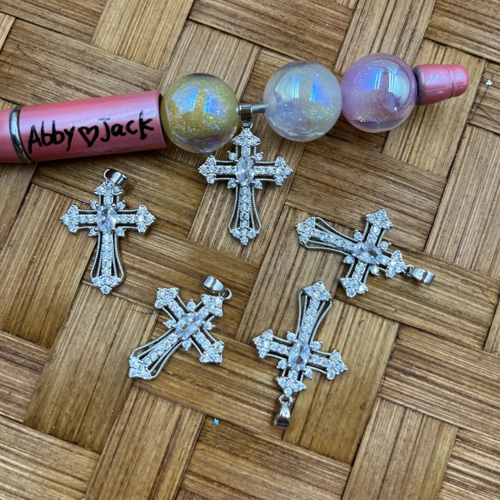 Four silver crystal rhinestone cross charm pendants with CZ stones and marquise center gem displayed on wicker surface next to a beadable pen with iridescent globe beads.