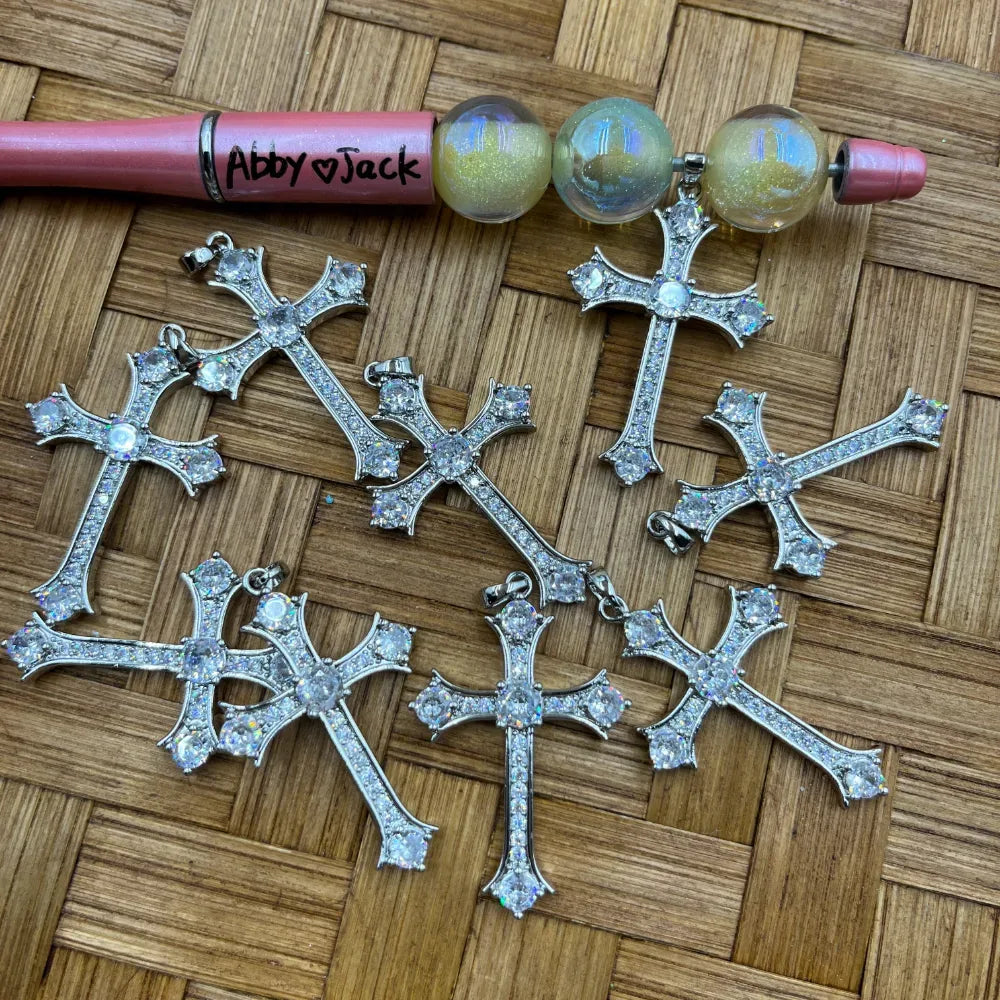 Six silver-tone crystal CZ rhinestone cross pendants with large center stone and pave detailing, displayed on a wicker surface next to iridescent globe bail beads and a pink beadable pen labeled Abby Jack.