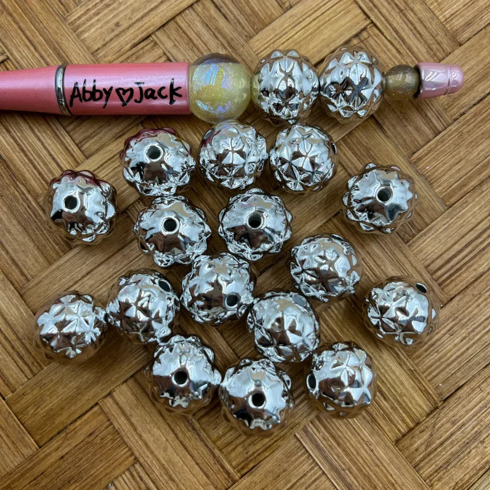 Shiny silver diamond-cut starburst patterned acrylic beads with metallic finish displayed on woven surface next to a beadable pen, showing hole detail for DIY jewelry and craft projects.