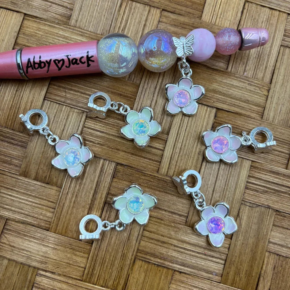 Silver flower charm bail beads with iridescent opal center stones in white and pink enamel, displayed on wicker surface next to a beadable pen with glitter globe beads.